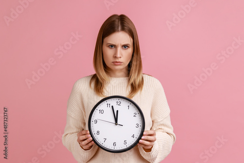 Portrait of strict bossy blond woman afraid of being late, holding in hand wall watch, deadline, punctuality, wearing white sweater. Indoor studio shot isolated on pink background.