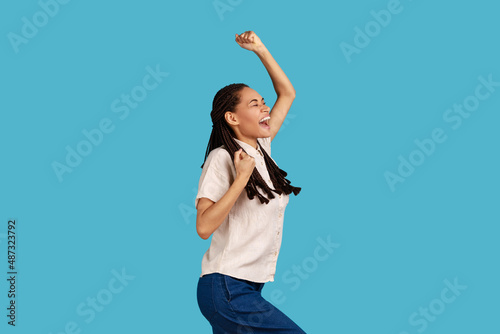 Portrait of excited woman with black dreadlocks clenches fists, celebrates success, feels happy after winning or triumph, wearing white shirt. Indoor studio shot isolated on blue background.
