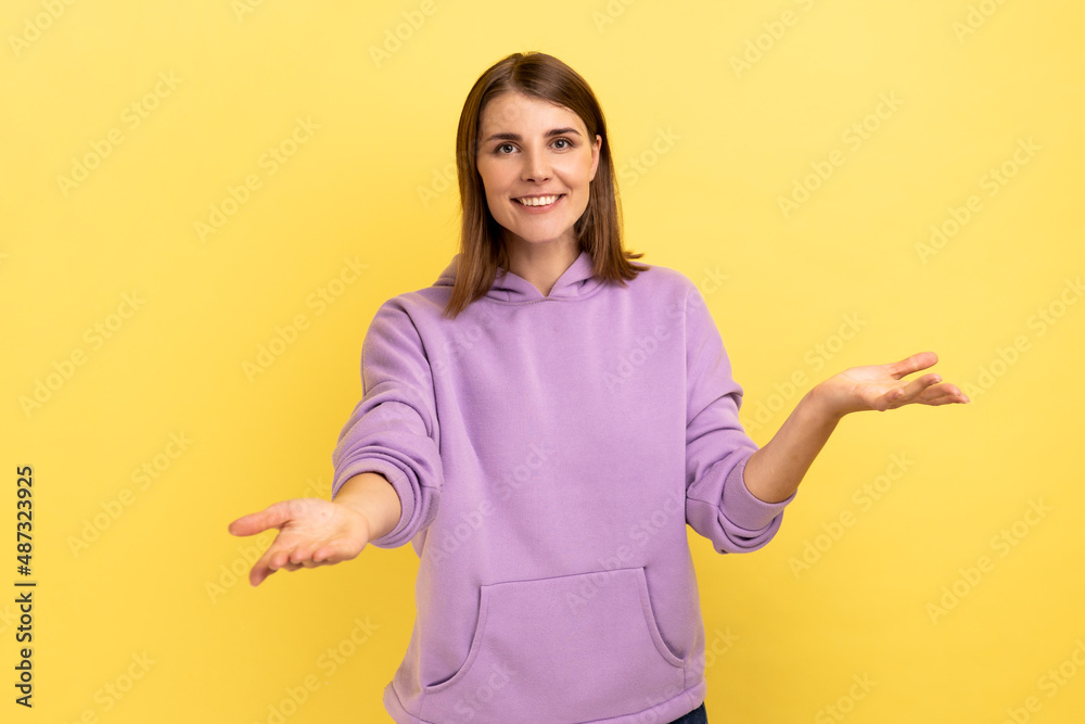 Openhearted generous woman outstretching hands looking at camera with kind smile, greeting and regaling, happy glad to see you, wearing purple hoodie. Indoor studio shot isolated on yellow background. Openhearted generous woman outstretching hands looking at camera with kind smile, greeting and regaling, happy glad to see you, wearing purple hoodie. Indoor studio shot isolated on yellow background.