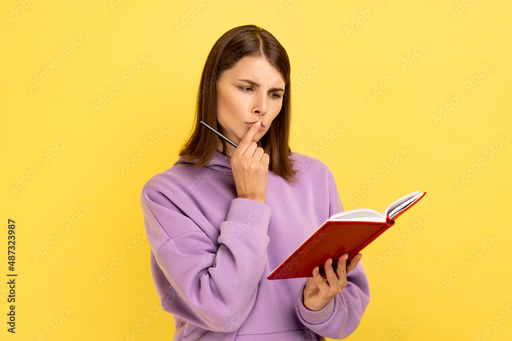 Thoughtful woman standing with paper notebook and pen, looking away with pensive expression, making to do list, wearing purple hoodie. Indoor studio shot isolated on yellow background. Thoughtful woman standing with paper notebook and pen, looking away with pensive expression, making to do list, wearing purple hoodie. Indoor studio shot isolated on yellow background.