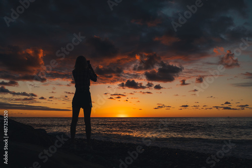 Silhouette of a young slender woman standing on the seashore and shooting a beautiful  colorful  magical sunset on a mobile phone