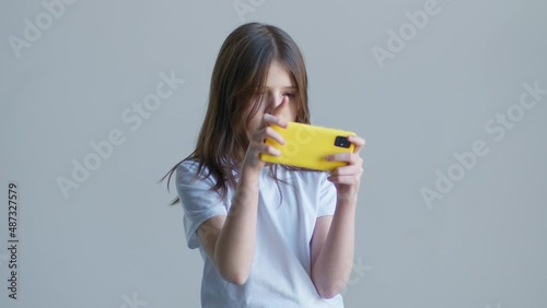 A blonde teenager girl in a white T-shirt game in the phone against the background of a white wall