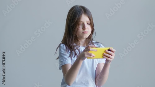 A blonde teenager girl in a white T-shirt game in the phone against the background of a white wall