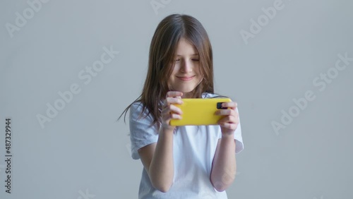 A blonde teenager girl in a white T-shirt looks at the phone and smiles against the background of a white wall