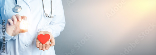 The doctor holds a red heart in one hand and an ampoule with a transparent liquid in the other (medicine, vaccine). Medical background with copy space and sunbeams. The concept of health, treatment, c