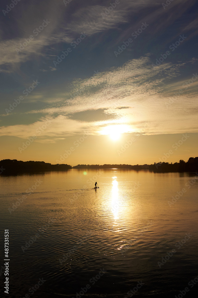 Sporty woman on paddle board at quiet river near ship with colorful sunset or sunrise