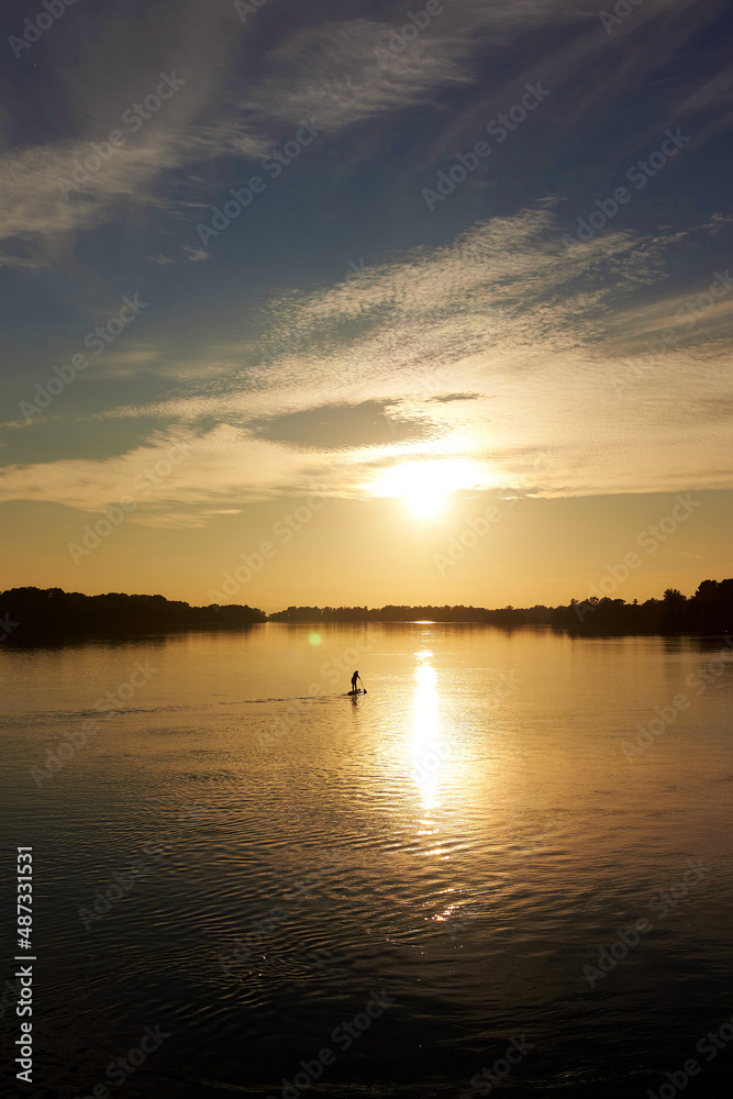 Sporty woman on paddle board at quiet river near ship with colorful sunset or sunrise Sporty woman on paddle board at quiet river near ship with colorful sunset or sunrise
