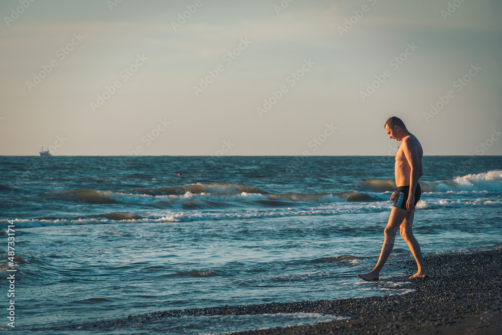 Happy man in swimming trunks stands on the seashore before sunset and enjoys life. Photo in movie style. Holiday by the sea Happy man in swimming trunks stands on the seashore before sunset and enjoys life. Photo in movie style. Holiday by the sea