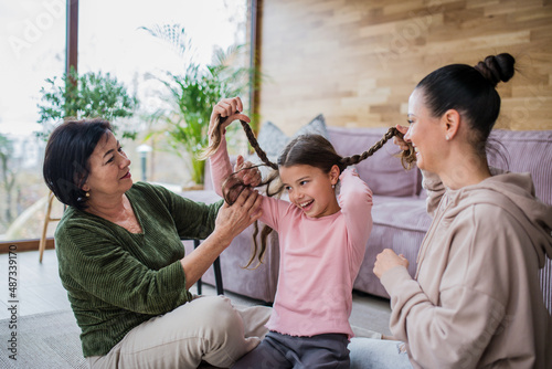 Canvas-taulu Happy little girl sitting on floor, her mother and grandmother making her plaits at home