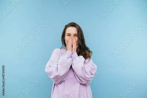 Portrait of a joyful teen girl in a sweatshirt on a blue background, happy looking at the camera and covering her face with her hands.