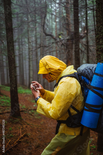 A male hiker on a hike through a dark misty forest in the rain uses a navigator on a smartphone to find his way.