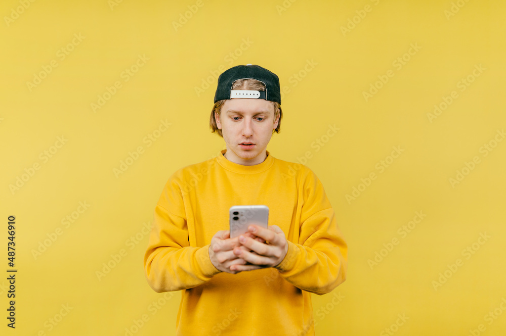 Handsome guy in a cap uses a smartphone with a serious face on a yellow background. Isolated Handsome guy in a cap uses a smartphone with a serious face on a yellow background. Isolated