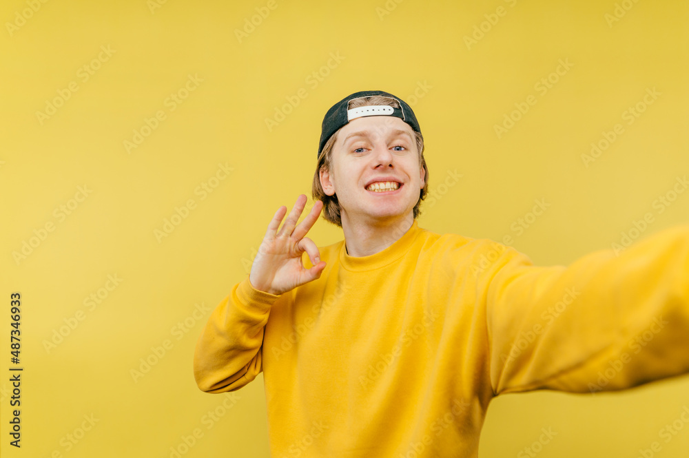 Joyful guy in a yellow sweatshirt and cap takes a selfie with a smile on his face and shows an OK gesture, isolated on a yellow background. Joyful guy in a yellow sweatshirt and cap takes a selfie with a smile on his face and shows an OK gesture, isolated on a yellow background.