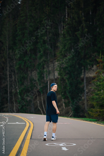 A young man in dark clothes walks on a footpath in the mountains against the backdrop of coniferous trees and looks away. Vertical