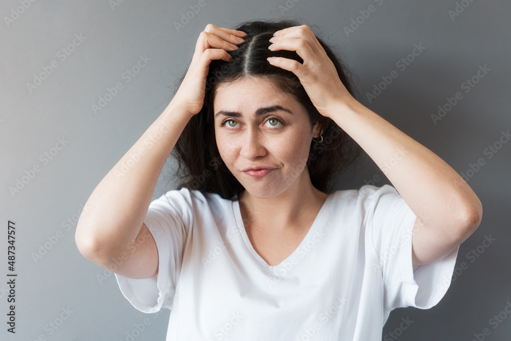 A young disgruntled woman looks up, touching the parting of her hair on her head with her hands. Gray background. The concept of hair problems and lices A young disgruntled woman looks up, touching the parting of her hair on her head with her hands. Gray background. The concept of hair problems and lices