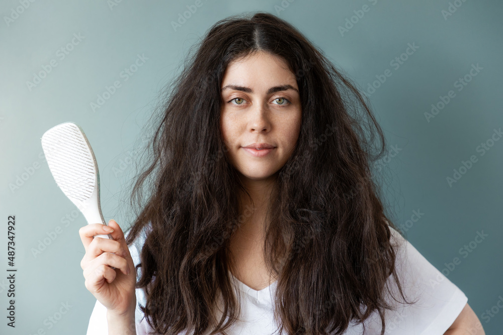 Portrait of a beautiful young woman with tangled brunette hear holding a comb. Blue background. The concept of hair care Portrait of a beautiful young woman with tangled brunette hear holding a comb. Blue background. The concept of hair care