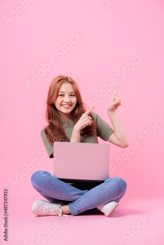 Young Asian woman sitting and using laptop on pink background