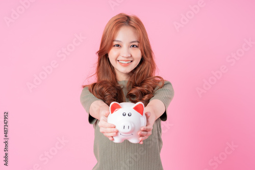 Young Asian woman holding piggy bank on pink background