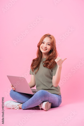 Young Asian woman sitting and using laptop on pink background © Timeimage