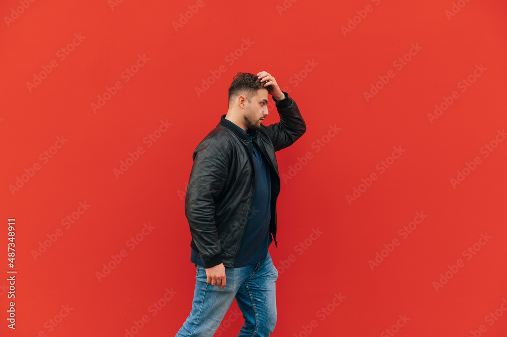 An adult man with a beard and a leather jacket walks against the backdrop of a red wall, side view. An adult man with a beard and a leather jacket walks against the backdrop of a red wall, side view.