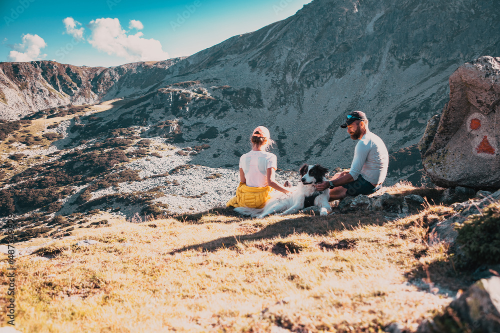 Fototapeta premium couple with dog sitting on rocks admiring mountain landscape slow travel