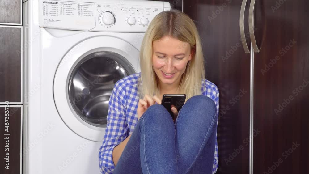 a happy modern woman using a home cleaning app on a smartphone in a modern home. She communicates in a messenger sitting by washing machine on floor. Bottles with the product are in foreground. a happy modern woman using a home cleaning app on a smartphone in a modern home. She communicates in a messenger sitting by washing machine on floor. Bottles with the product are in foreground.