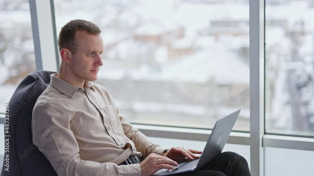 Caucasian male sitting in armchair and holding a laptop on his knees. Working man at the backdrop of panoramic window. Caucasian male sitting in armchair and holding a laptop on his knees. Working man at the backdrop of panoramic window.