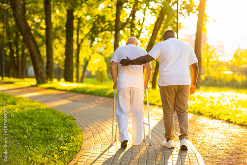 Caregiver is teaching old man to walk with walker. Professional nurse and patient walking outdoor at sunset. Assistance, rehabilitation and health care. photo