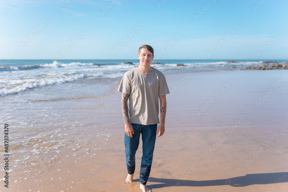 young caucasian man with full-arm tattoo in casual clothing walking on the beach