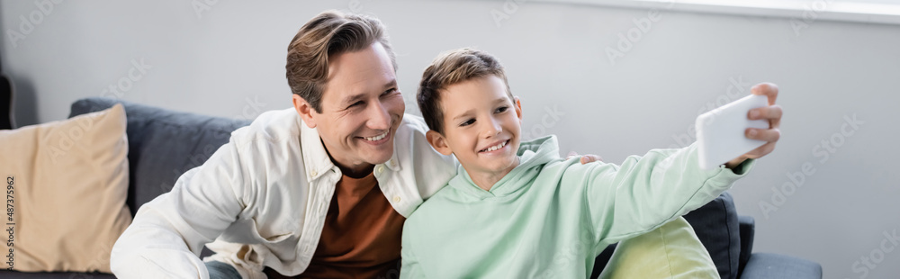 Positive parent and preteen son taking selfie on smartphone at home, banner Positive parent and preteen son taking selfie on smartphone at home, banner