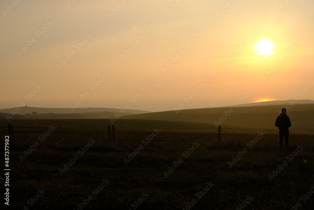 Eastbourne, East Sussex, United Kingdom - 9 October 2021. Man alone standing on hillside looking out across farmland towards a setting sun Copy space for label text banner or advertisement. Eastbourne, East Sussex, United Kingdom - 9 October 2021. Man alone standing on hillside looking out across farmland towards a setting sun Copy space for label text banner or advertisement.