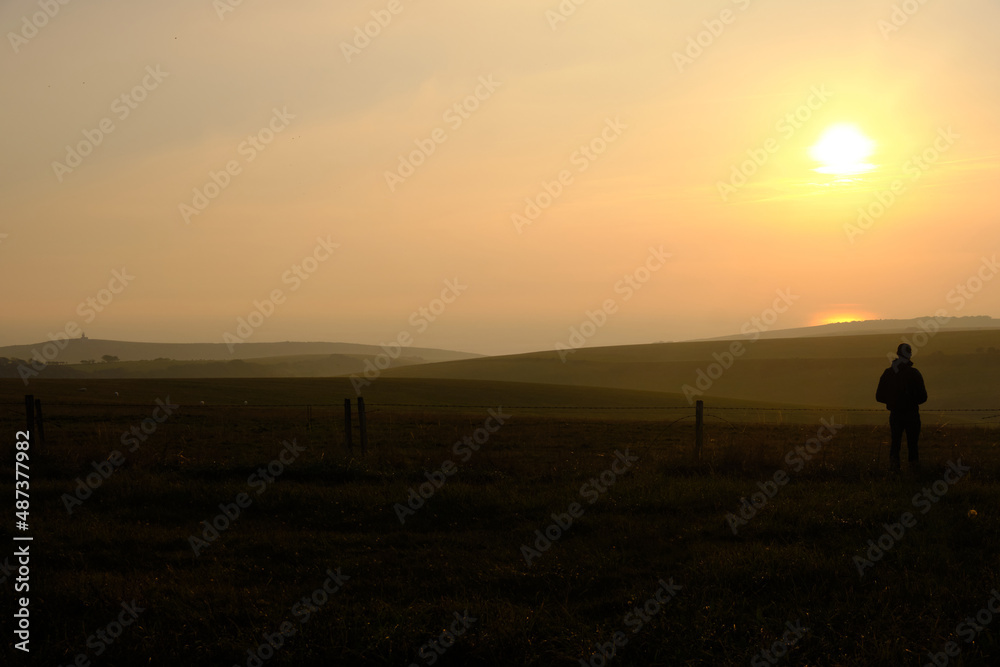 Eastbourne, East Sussex, United Kingdom - 9 October 2021. Man alone standing on hillside looking out across farmland towards a setting sun Copy space for label text banner or advertisement.