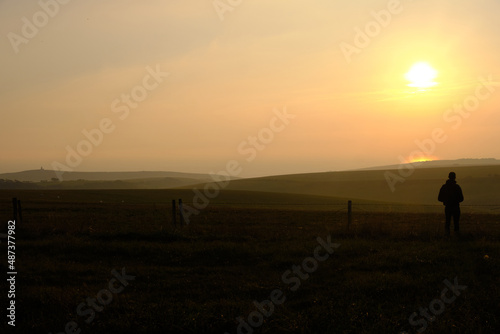 Eastbourne, East Sussex, United Kingdom - 9 October 2021. Man alone standing on hillside looking out across farmland towards a setting sun Copy space for label text banner or advertisement.