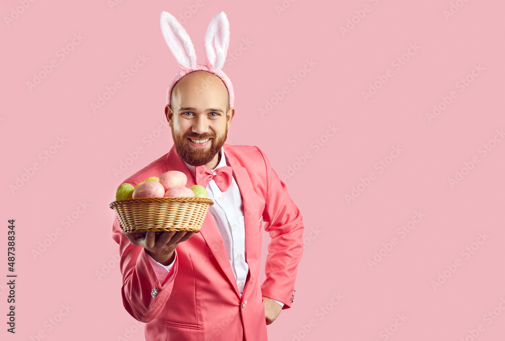 Portrait of happy funny handsome bearded young man in pink suit, bow tie and cute bunny ears standing isolated on color background, looking at camera, showing wicker bowl with Easter eggs and smiling Portrait of happy funny handsome bearded young man in pink suit, bow tie and cute bunny ears standing isolated on color background, looking at camera, showing wicker bowl with Easter eggs and smiling