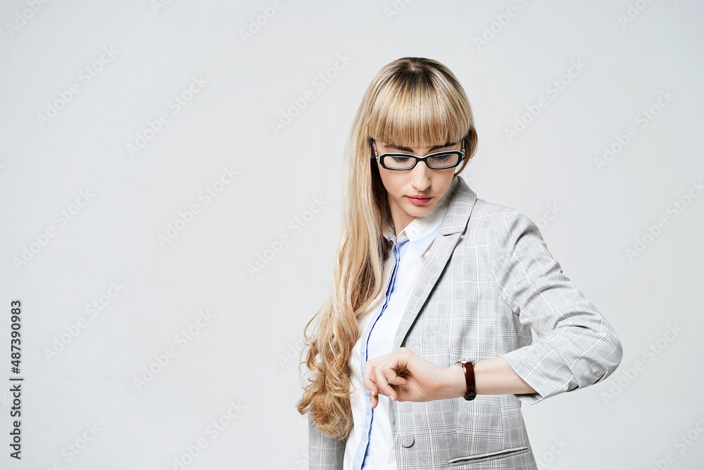 Young beautiful blonde woman in studio on light gray background. Model in pantsuit and glasses checking time with a wristwatch.