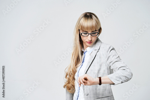 Obraz na płótnie Young beautiful blonde woman in studio on light gray background. Model in pantsuit and glasses checking time with a wristwatch. w salonie