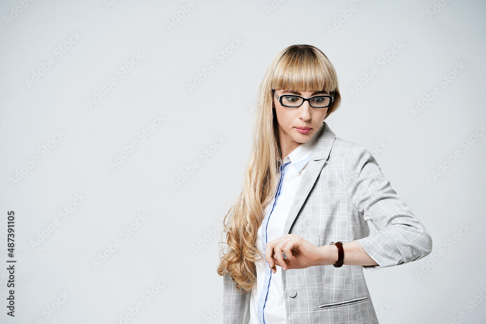 Young beautiful blonde woman in studio on light gray background. Model in pantsuit and glasses checking time with a wristwatch. Concept of being late.