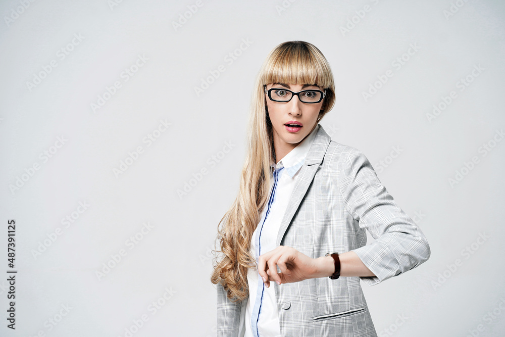 Young beautiful blonde woman in studio on light gray background. Model in pantsuit and glasses checking time with a wristwatch. Dissatisfied manager.