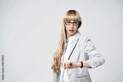 Obraz na płótnie Young beautiful blonde woman in studio on light gray background. Model in pantsuit and glasses checking time with a wristwatch. Dissatisfied manager. w salonie