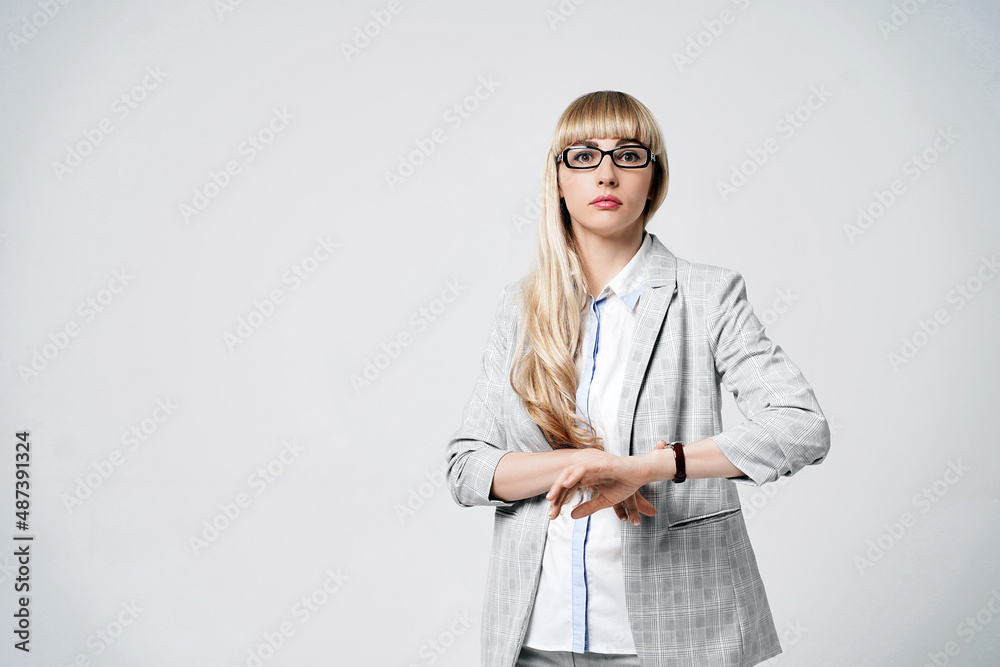 Young beautiful blonde woman in studio on light gray background. Model in pantsuit and glasses checking time with a wristwatch. Dissatisfied manager.