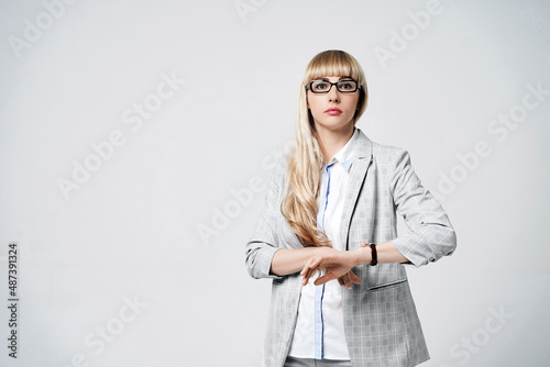 Obraz na płótnie Young beautiful blonde woman in studio on light gray background. Model in pantsuit and glasses checking time with a wristwatch. Dissatisfied manager. w salonie