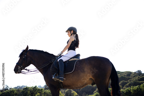 Portrait of a beautiful happy white woman smiling in her competition uniform and wearing a helmet while riding her Spanish bay horse on a sunny day with a forest in the background