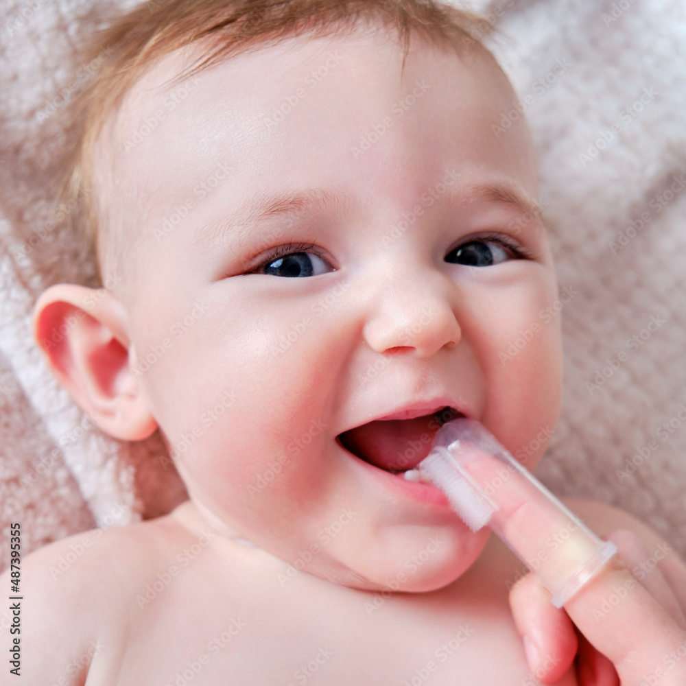 Mother hands brushing teeth with a finger brush of a happy infant baby. Mom doing oral hygiene to a smiling toddler kid, six to seven months old Mother hands brushing teeth with a finger brush of a happy infant baby. Mom doing oral hygiene to a smiling toddler kid, six to seven months old