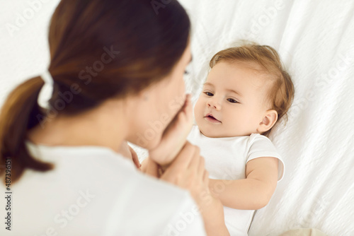 Happy mother and her cute barefoot child cuddling in cozy, warm bed together. Young mom tenderly holding and kissing her baby boy's soft little feet. Family, motherhood, love, and care concepts