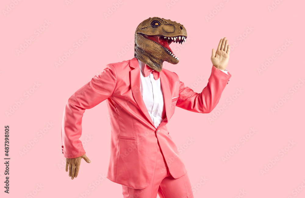 Funny man in rubber dinosaur mask dancing and having fun in the studio. Happy lizard headed guy in stylish funky vibrant pink party suit doing Egyptian dance moves isolated on pink colour background - obrazy, fototapety, plakaty 