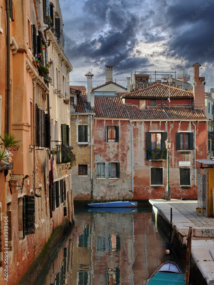 Hermoso rincón de Venecia sobre el río San Pantaleón con paredes color naranja que se reflejan sobre el agua y dos embarcaciones. Un cielo de nubes oscuras deja pasar unos rayos de sol.