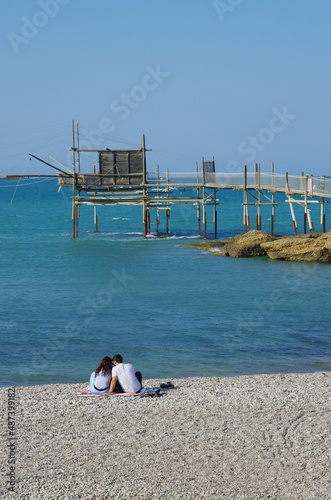 Punta Aderci Nature Reserve - Costa dei trabocchi - Abruzzo - A couple sitting on a pebble beach relaxes looking at the sea. In the background the imposing trabucco an ancient fishing machine. photo