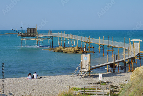 Punta Aderci Nature Reserve - Costa dei trabocchi - Abruzzo - A couple sitting on a pebble beach relaxes looking at the sea. In the background the imposing trabucco an ancient fishing machine.