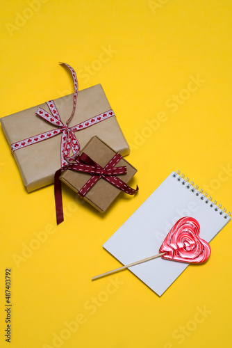 on a yellow background, a red heart-shaped lollipop, two gift boxes, a notepad. card, congratulations on Valentine's Day. romantic background
