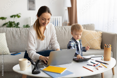 Freelancing at maternity leave. Young mother working on laptop while her toddler son playing with calculator neaby photo